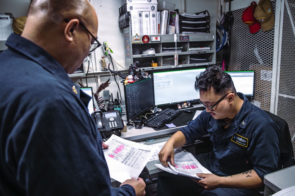 Two military personnel in dark navy uniforms are reviewing printed documents with highlighted entries in what appears to be an administrative or operations office aboard a naval vessel. The seated individual has a name tag visible on their uniform. The workspace is compact and well-equipped with computers, a desk phone, radios, binders, and various personal items.