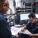 Two military personnel in dark navy uniforms are reviewing printed documents with highlighted entries in what appears to be an administrative or operations office aboard a naval vessel. The seated individual has a name tag visible on their uniform. The workspace is compact and well-equipped with computers, a desk phone, radios, binders, and various personal items.
