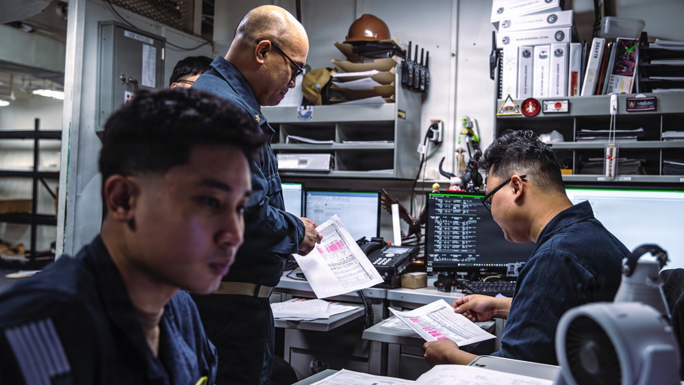 Three military personnel in dark navy uniforms are working inside what appears to be a shipboard operations or administrative office. They are reviewing printed documents with pink-highlighted data, surrounded by computer monitors, radios, binders, and various equipment. The setting is consistent with a U.S. Navy vessel's interior workspace.