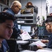 Three military personnel in dark navy uniforms are working inside what appears to be a shipboard operations or administrative office. They are reviewing printed documents with pink-highlighted data, surrounded by computer monitors, radios, binders, and various equipment. The setting is consistent with a U.S. Navy vessel's interior workspace.