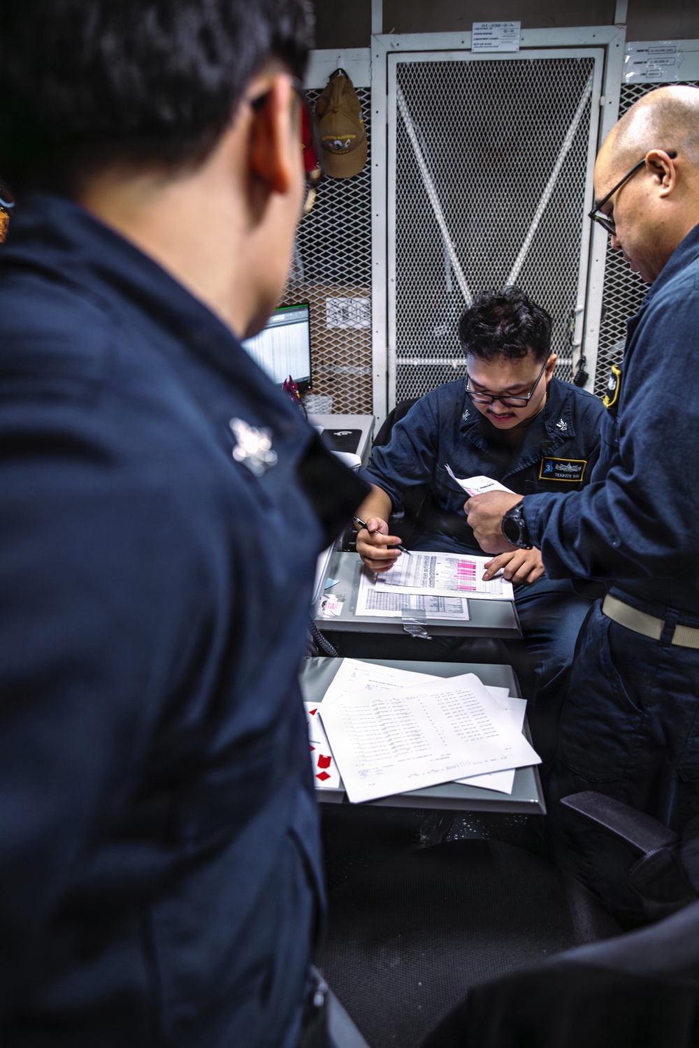 Three individuals wearing dark navy blue uniforms are gathered around a desk reviewing documents and paperwork. The central figure, wearing glasses and a name tag, appears to be writing or signing documents while a colleague points at the paperwork. The setting appears to be a military or naval workspace with a mesh storage cage visible in the background.