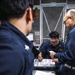Three individuals wearing dark navy blue uniforms are gathered around a desk reviewing documents and paperwork. The central figure, wearing glasses and a name tag, appears to be writing or signing documents while a colleague points at the paperwork. The setting appears to be a military or naval workspace with a mesh storage cage visible in the background.