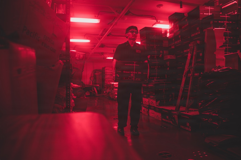 A person is standing in a storage room or warehouse bathed in red lighting, carrying a box of Hammermill 'Great White 30' copy paper. The space is densely packed with stacked boxes of the same paper brand on shelving units. The red-tinted fluorescent lighting creates a dramatic, moody atmosphere throughout the storage area.