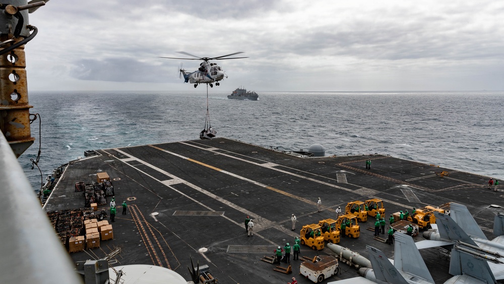 A military helicopter is conducting a vertical replenishment (VERTREP) operation over the flight deck of a large naval vessel, likely an aircraft carrier or amphibious assault ship. The helicopter is carrying a slung load while crew members in green jerseys and yellow forklifts stand by on the deck to receive and distribute supplies. A support or replenishment ship is visible in the background on the open ocean.