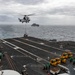 A military helicopter is conducting a vertical replenishment (VERTREP) operation over the flight deck of a large naval vessel, likely an aircraft carrier or amphibious assault ship. The helicopter is carrying a slung load while crew members in green jerseys and yellow forklifts stand by on the deck to receive and distribute supplies. A support or replenishment ship is visible in the background on the open ocean.