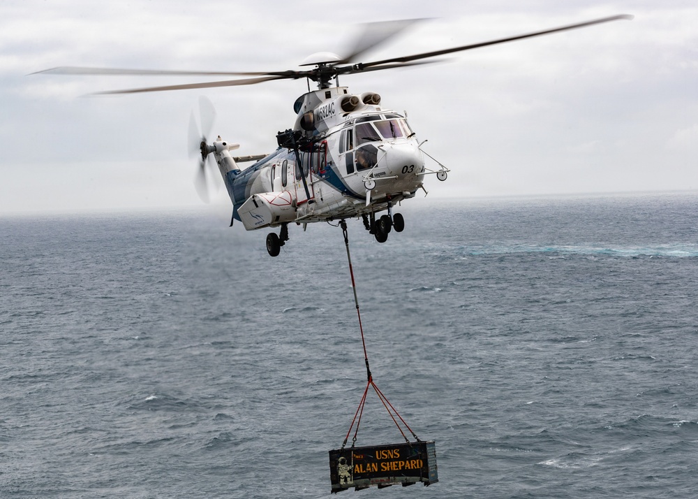 A white civilian-style helicopter marked '03' with registration N682AC is performing a vertical replenishment (VERTREP) operation over open ocean. It is carrying a cargo net sling load bearing a sign that reads 'USNS ALAN SHEPARD', indicating the load originates from or is destined for that U.S. Navy logistics vessel. The overcast sky and choppy sea conditions suggest an active maritime environment.