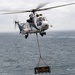 A white civilian-style helicopter marked '03' with registration N682AC is performing a vertical replenishment (VERTREP) operation over open ocean. It is carrying a cargo net sling load bearing a sign that reads 'USNS ALAN SHEPARD', indicating the load originates from or is destined for that U.S. Navy logistics vessel. The overcast sky and choppy sea conditions suggest an active maritime environment.