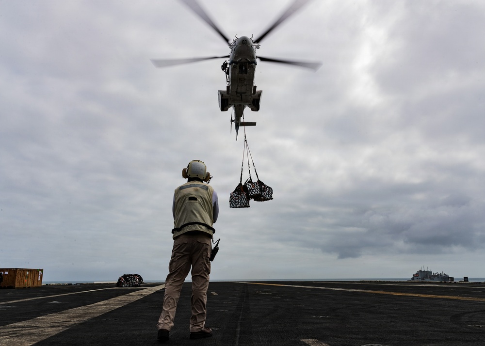 A military helicopter hovers low over a naval vessel's flight deck while conducting a vertical replenishment (VERTREP) operation, delivering cargo suspended in cargo nets. A deck crew member wearing a helmet and safety vest stands on the flight deck, guiding the helicopter and its suspended load. A second ship is visible on the horizon in the background.