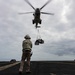 A military helicopter hovers low over a naval vessel's flight deck while conducting a vertical replenishment (VERTREP) operation, delivering cargo suspended in cargo nets. A deck crew member wearing a helmet and safety vest stands on the flight deck, guiding the helicopter and its suspended load. A second ship is visible on the horizon in the background.