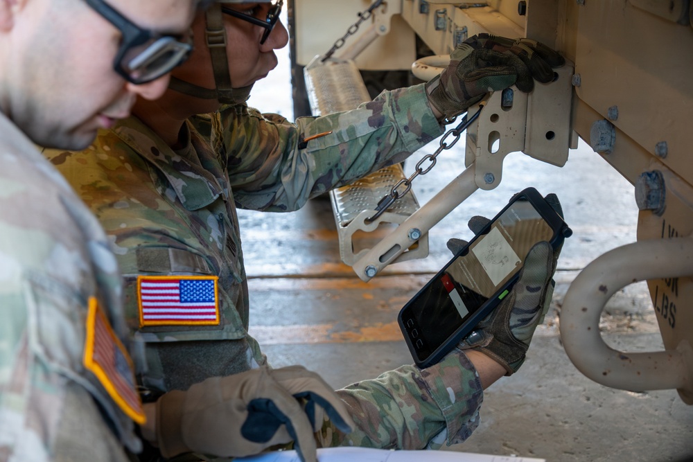 Two U.S. Army soldiers in Operational Camouflage Pattern (OCP) uniforms are conducting an inspection or maintenance check on a tan military vehicle. One soldier is holding a tablet device, likely scanning or recording data, while the other examines a mechanical component involving a chain and bracket assembly. The American flag patch is clearly visible on one soldier's left shoulder.