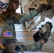 Two U.S. Army soldiers in Operational Camouflage Pattern (OCP) uniforms are conducting an inspection or maintenance check on a tan military vehicle. One soldier is holding a tablet device, likely scanning or recording data, while the other examines a mechanical component involving a chain and bracket assembly. The American flag patch is clearly visible on one soldier's left shoulder.