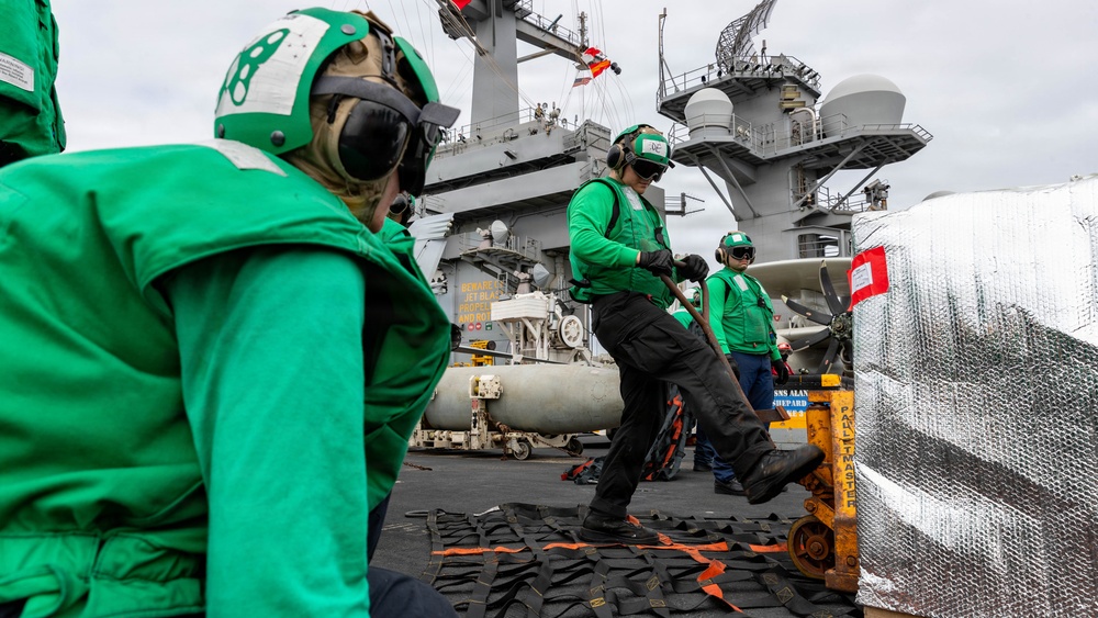 The image shows U.S. Navy personnel in green jerseys working on the flight deck of an aircraft carrier, handling cargo or equipment using a yellow pallet jack. The workers are wearing standard flight deck safety gear including cranials (helmets), goggles, and hearing protection. A large reflective/insulated pallet wrapped in metallic material is visible in the foreground, with the ship's island superstructure visible in the background.