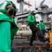 The image shows U.S. Navy personnel in green jerseys working on the flight deck of an aircraft carrier, handling cargo or equipment using a yellow pallet jack. The workers are wearing standard flight deck safety gear including cranials (helmets), goggles, and hearing protection. A large reflective/insulated pallet wrapped in metallic material is visible in the foreground, with the ship's island superstructure visible in the background.