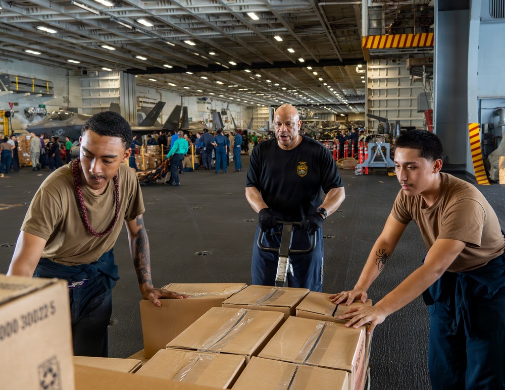 Three individuals are working together to move cardboard boxes on a pallet jack inside what appears to be the hangar bay of a U.S. Navy aircraft carrier. Military aircraft, including what appear to be F-35s, are visible in the background along with numerous personnel engaged in logistics operations. The scene depicts a replenishment or cargo-handling evolution typical of carrier operations.