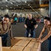 Three individuals are working together to move cardboard boxes on a pallet jack inside what appears to be the hangar bay of a U.S. Navy aircraft carrier. Military aircraft, including what appear to be F-35s, are visible in the background along with numerous personnel engaged in logistics operations. The scene depicts a replenishment or cargo-handling evolution typical of carrier operations.