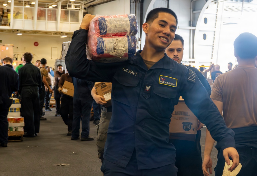 A U.S. Navy sailor identified by name tag as 'NOE CASTELO' carries packaged supplies on his shoulder while smiling during what appears to be a resupply or logistics operation aboard a naval vessel. Multiple personnel are visible in the background handling boxes and cargo in what looks like a hangar bay or cargo hold. The scene depicts a coordinated supply chain effort with numerous service members working together.