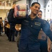 A U.S. Navy sailor identified by name tag as 'NOE CASTELO' carries packaged supplies on his shoulder while smiling during what appears to be a resupply or logistics operation aboard a naval vessel. Multiple personnel are visible in the background handling boxes and cargo in what looks like a hangar bay or cargo hold. The scene depicts a coordinated supply chain effort with numerous service members working together.