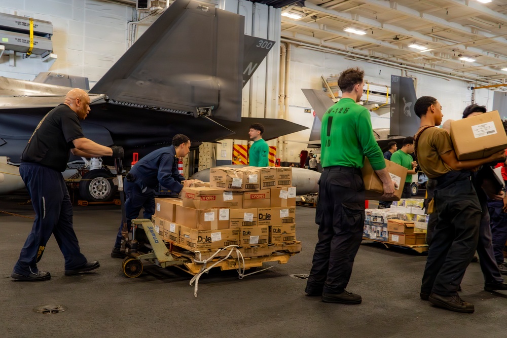 Military personnel are conducting a resupply operation inside what appears to be an aircraft carrier hangar bay. Sailors and crew members are moving pallets and boxes of food supplies, including visible Dole and Lyons branded products, using a pallet jack. At least two military aircraft, appearing to be F-35s based on their stealth profile, are parked in the background.