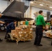 Military personnel are conducting a resupply operation inside what appears to be an aircraft carrier hangar bay. Sailors and crew members are moving pallets and boxes of food supplies, including visible Dole and Lyons branded products, using a pallet jack. At least two military aircraft, appearing to be F-35s based on their stealth profile, are parked in the background.