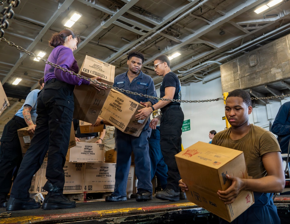 U.S. Navy personnel are conducting a stores replenishment operation aboard what appears to be a naval vessel, passing cardboard boxes in a chain-gang style supply line. The boxes are visibly labeled as Kellogg's Favorite Variety Pack food supplies marked 'FS' (Food Service). The setting is an interior hangar or cargo bay of a ship, identifiable by the industrial ceiling, chains, and military-style flooring.