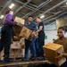 U.S. Navy personnel are conducting a stores replenishment operation aboard what appears to be a naval vessel, passing cardboard boxes in a chain-gang style supply line. The boxes are visibly labeled as Kellogg's Favorite Variety Pack food supplies marked 'FS' (Food Service). The setting is an interior hangar or cargo bay of a ship, identifiable by the industrial ceiling, chains, and military-style flooring.
