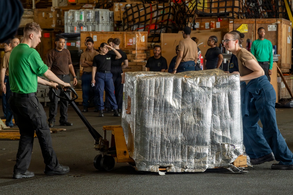 Military or naval personnel are moving a large pallet wrapped in reflective thermal/mylar insulation using a manual pallet jack in what appears to be a ship's cargo hold or hangar bay. Two workers in the foreground are actively maneuvering the pallet — one pulling the pallet jack and one pushing the load — while a group of approximately 15 observers stand in the background. The setting is consistent with a U.S. Navy vessel's interior storage or logistics area.