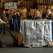 Military or naval personnel are moving a large pallet wrapped in reflective thermal/mylar insulation using a manual pallet jack in what appears to be a ship's cargo hold or hangar bay. Two workers in the foreground are actively maneuvering the pallet — one pulling the pallet jack and one pushing the load — while a group of approximately 15 observers stand in the background. The setting is consistent with a U.S. Navy vessel's interior storage or logistics area.