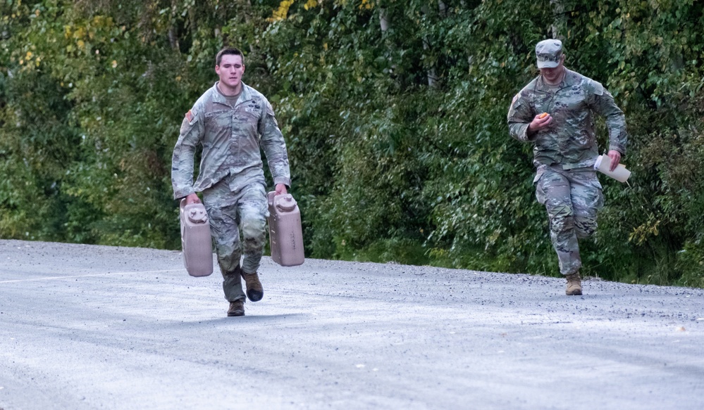 Two soldiers in US Army Operational Camouflage Pattern (OCP) uniforms are moving along a gravel path bordered by dense green trees. The soldier in the foreground is carrying two large military-style water or fuel jerry cans, one in each hand, while the soldier behind carries a single container. Both appear to be in motion, suggesting a training exercise or logistical task.