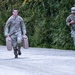 Two soldiers in US Army Operational Camouflage Pattern (OCP) uniforms are moving along a gravel path bordered by dense green trees. The soldier in the foreground is carrying two large military-style water or fuel jerry cans, one in each hand, while the soldier behind carries a single container. Both appear to be in motion, suggesting a training exercise or logistical task.