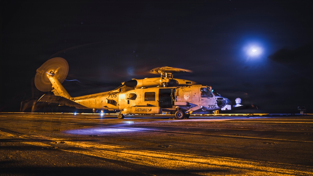 A U.S. Navy MH-60R Seahawk helicopter, bureau number 705, assigned to the 'Saberhawks' squadron, is parked on what appears to be a naval vessel flight deck at night. The helicopter is illuminated by warm artificial lighting, with rotor blades appearing to be spinning or recently stopped. A bright blue light source, likely a spotlight or floodlight, is visible in the background.