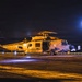 A U.S. Navy MH-60R Seahawk helicopter, bureau number 705, assigned to the 'Saberhawks' squadron, is parked on what appears to be a naval vessel flight deck at night. The helicopter is illuminated by warm artificial lighting, with rotor blades appearing to be spinning or recently stopped. A bright blue light source, likely a spotlight or floodlight, is visible in the background.