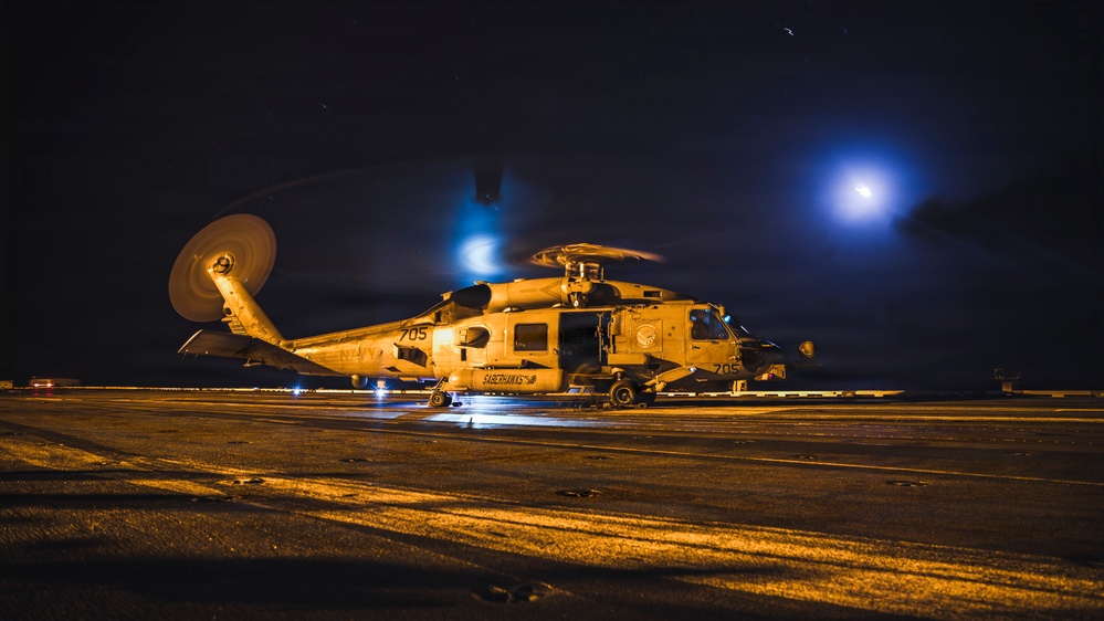A U.S. Navy helicopter, numbered 705 and belonging to the 'Saberhawks' squadron, is photographed on what appears to be a ship's flight deck at night. The rotors are spinning, indicated by motion blur, and the scene is dramatically lit by warm orange deck lighting and a bright blue light source in the background. The overall atmosphere suggests an active night flight operation aboard a naval vessel.