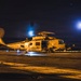 A U.S. Navy helicopter, numbered 705 and belonging to the 'Saberhawks' squadron, is photographed on what appears to be a ship's flight deck at night. The rotors are spinning, indicated by motion blur, and the scene is dramatically lit by warm orange deck lighting and a bright blue light source in the background. The overall atmosphere suggests an active night flight operation aboard a naval vessel.
