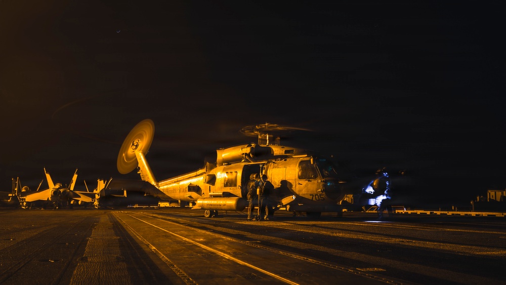 A nighttime scene on what appears to be a naval aircraft carrier flight deck, illuminated by orange/amber artificial lighting. A military helicopter, bearing the number '705', is the central subject with its rotors spinning, surrounded by ground crew personnel. Fixed-wing aircraft are visible in the background to the left.