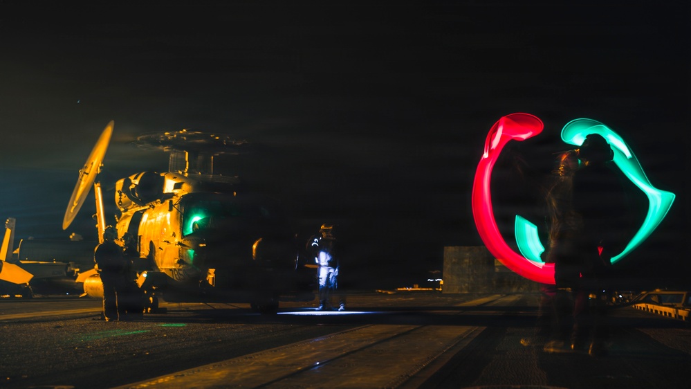 A nighttime military flight deck operation showing a helicopter on what appears to be a naval vessel or airfield. Personnel are conducting marshalling operations using illuminated wands, with the long-exposure photography capturing circular light trails in red and green. The scene depicts standard night flight deck crew procedures with multiple ground crew members visible.