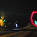 A nighttime military flight deck operation showing a helicopter on what appears to be a naval vessel or airfield. Personnel are conducting marshalling operations using illuminated wands, with the long-exposure photography capturing circular light trails in red and green. The scene depicts standard night flight deck crew procedures with multiple ground crew members visible.
