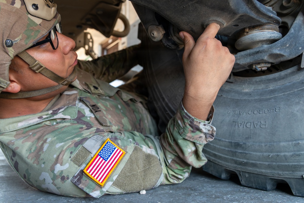 A U.S. Army soldier in Operational Camouflage Pattern (OCP) uniform is lying on the ground performing maintenance or inspection work on a military vehicle. The soldier is working near the wheel and undercarriage area of the vehicle, using their hands to manipulate a component. The image is a close-up shot emphasizing the soldier's focused effort on the mechanical task.