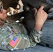 A U.S. Army soldier in Operational Camouflage Pattern (OCP) uniform is lying on the ground performing maintenance or inspection work on a military vehicle. The soldier is working near the wheel and undercarriage area of the vehicle, using their hands to manipulate a component. The image is a close-up shot emphasizing the soldier's focused effort on the mechanical task.