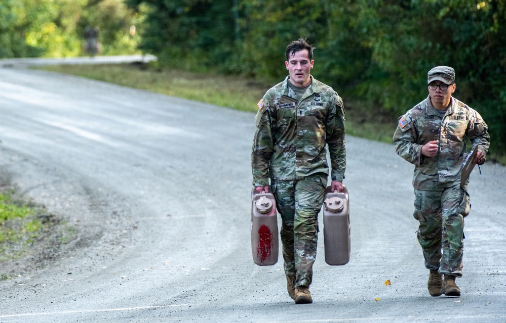 Two U.S. Army soldiers in MultiCam (OCP) camouflage uniforms are walking along a paved road flanked by dense green vegetation. The soldier in the foreground is carrying two large jerry cans, one of which has a red liquid stain on it, while the soldier behind appears to be holding a clipboard. This appears to be a physical fitness or training event, likely a stress test or competition.