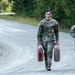 Two U.S. Army soldiers in MultiCam (OCP) camouflage uniforms are walking along a paved road flanked by dense green vegetation. The soldier in the foreground is carrying two large jerry cans, one of which has a red liquid stain on it, while the soldier behind appears to be holding a clipboard. This appears to be a physical fitness or training event, likely a stress test or competition.