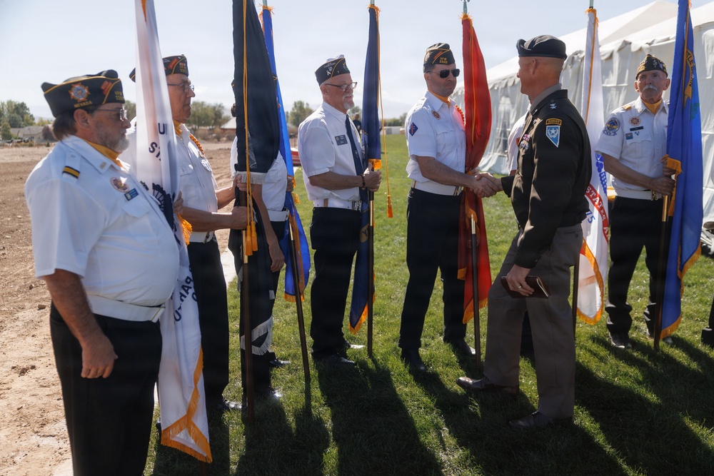 A color guard ceremony is taking place outdoors on a grassy area, featuring several veterans in white shirts and garrison caps holding various flags. A man in an olive/dark military-style uniform is shaking hands with one of the color guard members, suggesting an official greeting or recognition event. The flags include what appear to be U.S. state and military branch flags, and the participants are wearing insignia consistent with a veterans organization such as the American Legion or VFW.