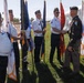 A color guard ceremony is taking place outdoors on a grassy area, featuring several veterans in white shirts and garrison caps holding various flags. A man in an olive/dark military-style uniform is shaking hands with one of the color guard members, suggesting an official greeting or recognition event. The flags include what appear to be U.S. state and military branch flags, and the participants are wearing insignia consistent with a veterans organization such as the American Legion or VFW.