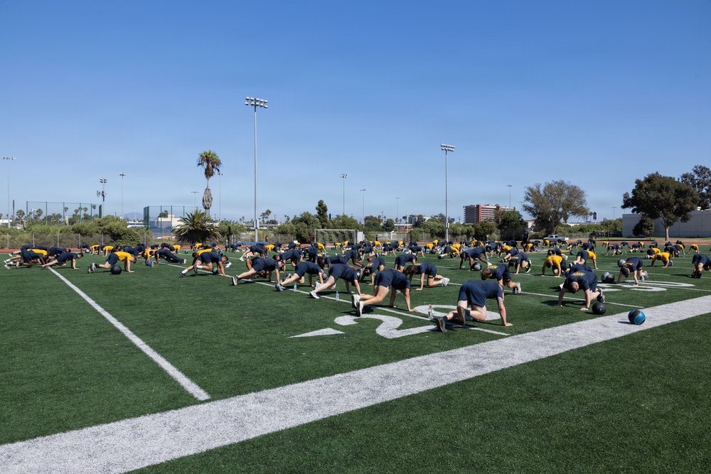 A large group of individuals dressed in matching navy blue and yellow athletic uniforms are performing synchronized stretching or exercise drills on an artificial turf football field. The group appears to be engaged in a structured physical training session, likely military or team-based given the uniformity of attire. The setting is an outdoor sports complex with stadium lights, palm trees, and urban buildings visible in the background under a clear blue sky.