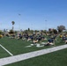 A large group of individuals dressed in matching navy blue and yellow athletic uniforms are performing synchronized stretching or exercise drills on an artificial turf football field. The group appears to be engaged in a structured physical training session, likely military or team-based given the uniformity of attire. The setting is an outdoor sports complex with stadium lights, palm trees, and urban buildings visible in the background under a clear blue sky.