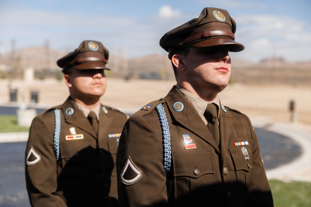 Two U.S. Army soldiers in dress uniforms stand at attention outdoors, likely during a formal ceremony or parade. Both wear olive drab Class A uniforms with garrison caps, rank insignia, and various awards and decorations. The setting appears to be a military installation in an arid or semi-arid region.