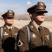 Two U.S. Army soldiers in dress uniforms stand at attention outdoors, likely during a formal ceremony or parade. Both wear olive drab Class A uniforms with garrison caps, rank insignia, and various awards and decorations. The setting appears to be a military installation in an arid or semi-arid region.