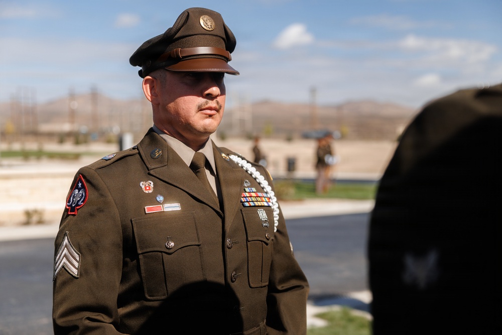 A U.S. Army soldier in Army Service Uniform (Class A) stands at attention outdoors, wearing a garrison cap with eagle insignia. The soldier displays multiple medals, ribbons, patches, and a white aiguillette, indicating a ceremonial or formal military event. The background shows an open outdoor area with mountains visible in the distance under a partly cloudy sky.
