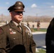 A U.S. Army soldier in Army Service Uniform (Class A) stands at attention outdoors, wearing a garrison cap with eagle insignia. The soldier displays multiple medals, ribbons, patches, and a white aiguillette, indicating a ceremonial or formal military event. The background shows an open outdoor area with mountains visible in the distance under a partly cloudy sky.