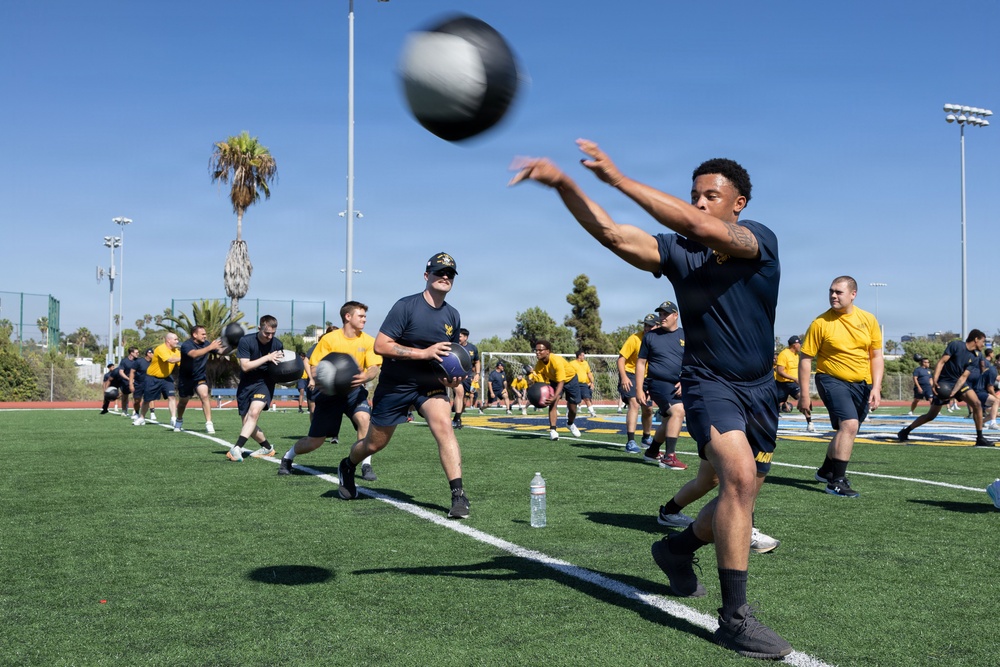 A large group of individuals in military-style uniforms (navy blue and yellow t-shirts with 'NAVY' markings) are conducting an outdoor physical training session on an artificial turf athletic field. Participants are performing medicine ball throwing exercises in pairs or lines across the field. The setting appears to be a sports complex in a warm, sunny climate, likely Southern California based on the palm trees and clear blue sky.