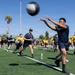 A large group of individuals in military-style uniforms (navy blue and yellow t-shirts with 'NAVY' markings) are conducting an outdoor physical training session on an artificial turf athletic field. Participants are performing medicine ball throwing exercises in pairs or lines across the field. The setting appears to be a sports complex in a warm, sunny climate, likely Southern California based on the palm trees and clear blue sky.